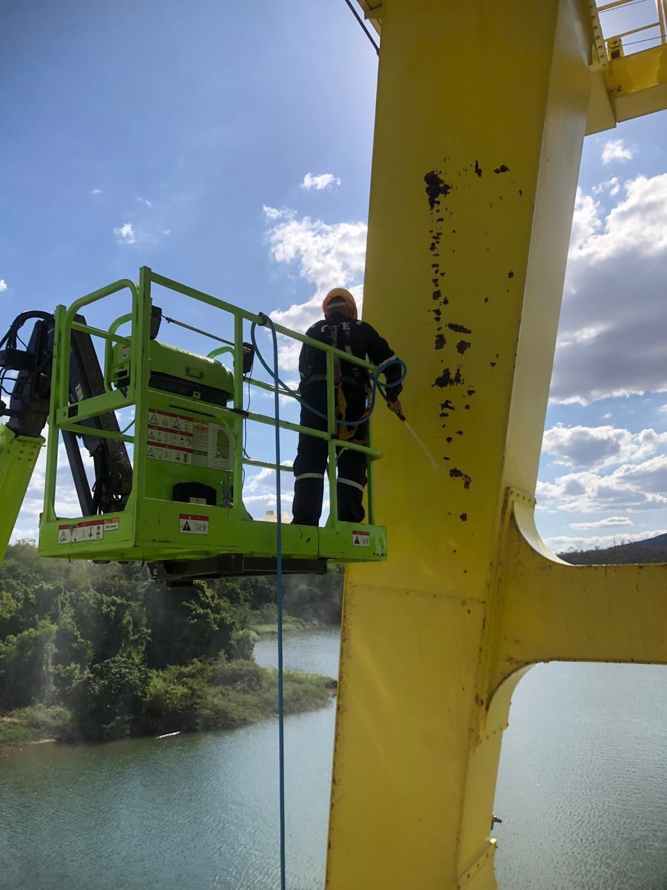 Técnico em cesto elevatório verde realizando jateamento de viga amarela sobre a represa, céu azul ao fundo