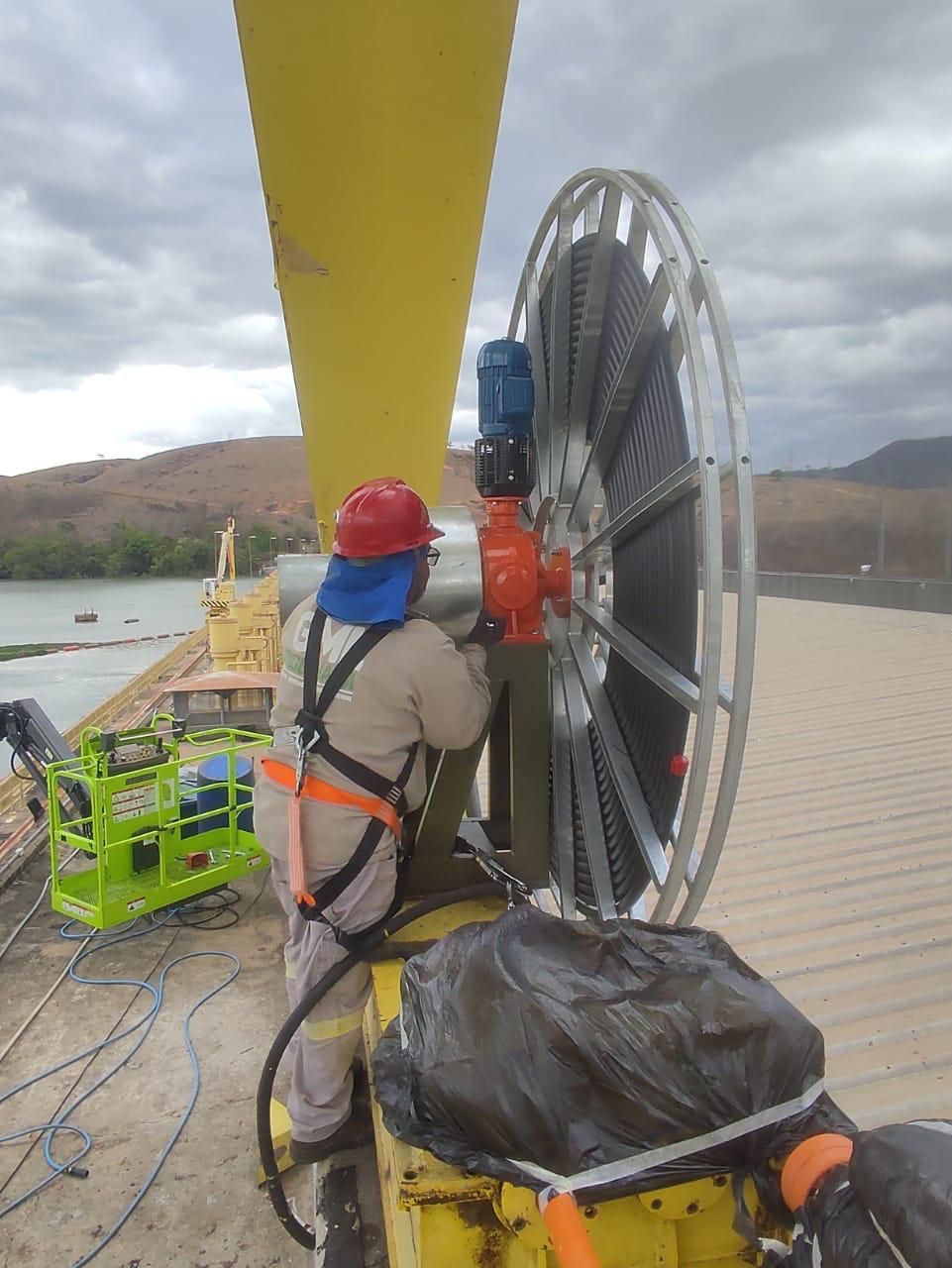 Técnico de capacete vermelho, com arnês completo, realizando manutenção em redutor sobre estrutura elevada de cor amarela, com uma represa e morros ao fundo