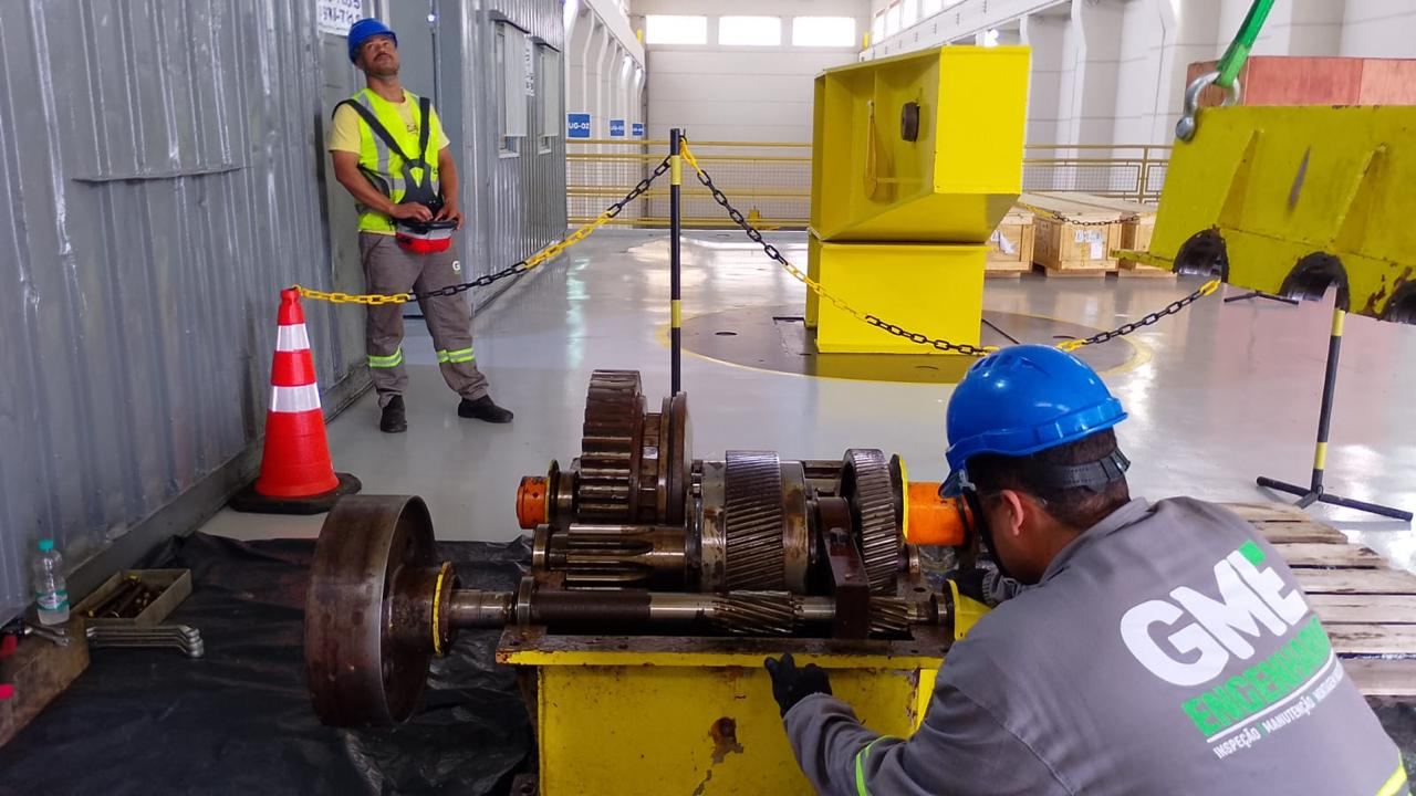 Dois técnicos inspecionando engrenagens expostas em usina, sinalizados por cones de segurança
