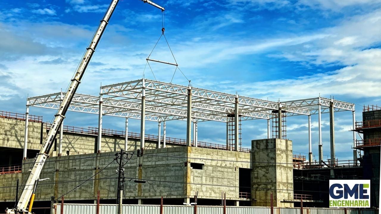 Dois técnicos em plataforma elevatória vermelha montando treliça metálica da cobertura, técnico em plataforma azul ao fundo