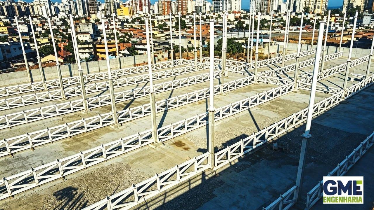 Treliça metálica sendo montada sobre torres de andaime, dois técnicos em plataformas elevadas, céu azul com nuvens