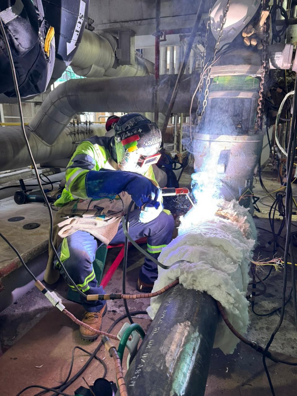 Soldador com máscara de proteção realizando soldagem em tubulação industrial, manta cerâmica de isolamento ao lado