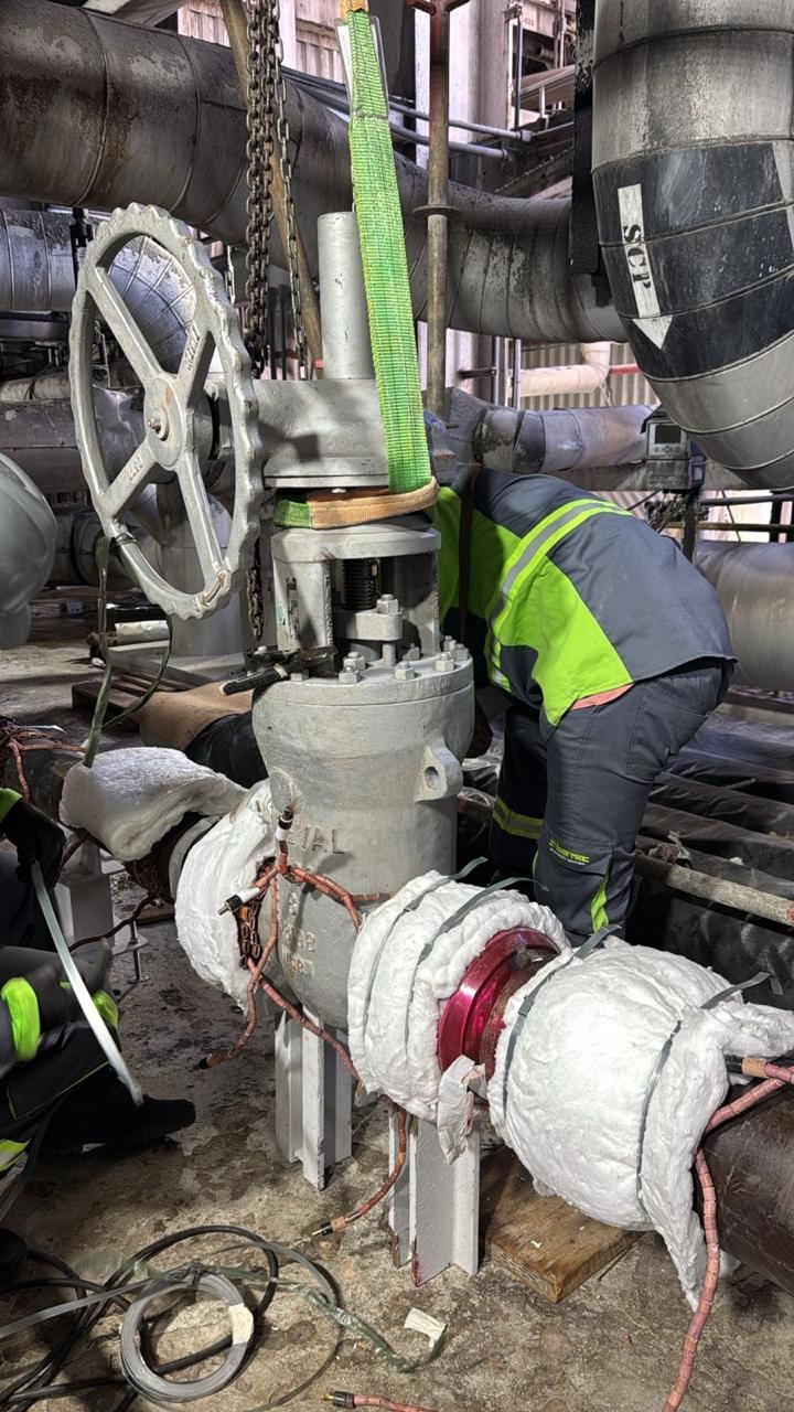 Técnico posicionando cinta de içamento verde em válvula industrial, isolamento térmico ao redor da tubulação