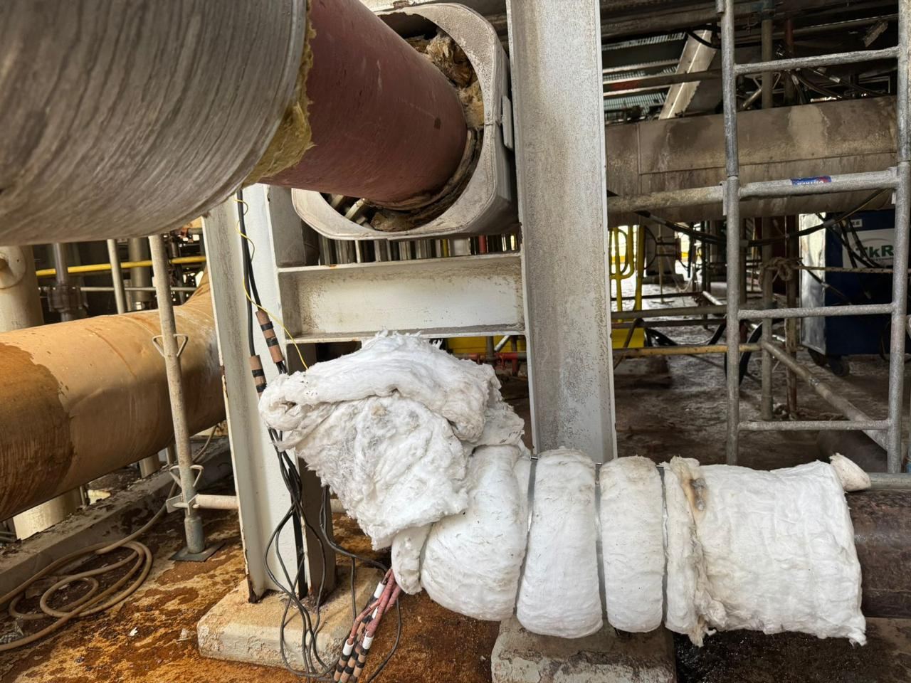 Tubulação industrial com manta de isolamento térmico envolta, estrutura metálica e passarela ao fundo