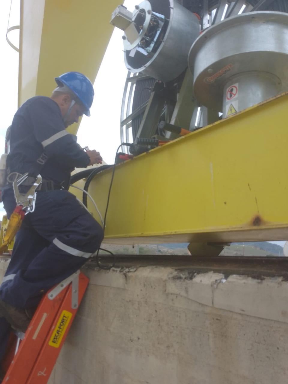 Redutor industrial sendo manuseado junto à viga amarela, represa ao fundo